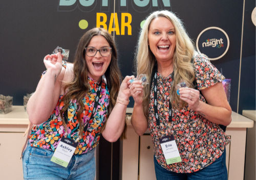 Two women smiling while holding small objects, standing in front of a sign that reads 'Button Bar.' They are wearing name badges.