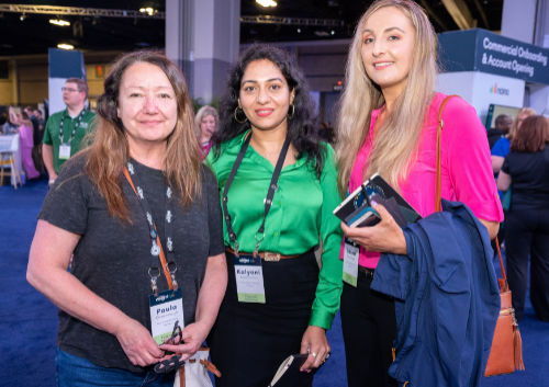 Three women standing together at an event, each wearing name tags. One holds a smartphone and a jacket.